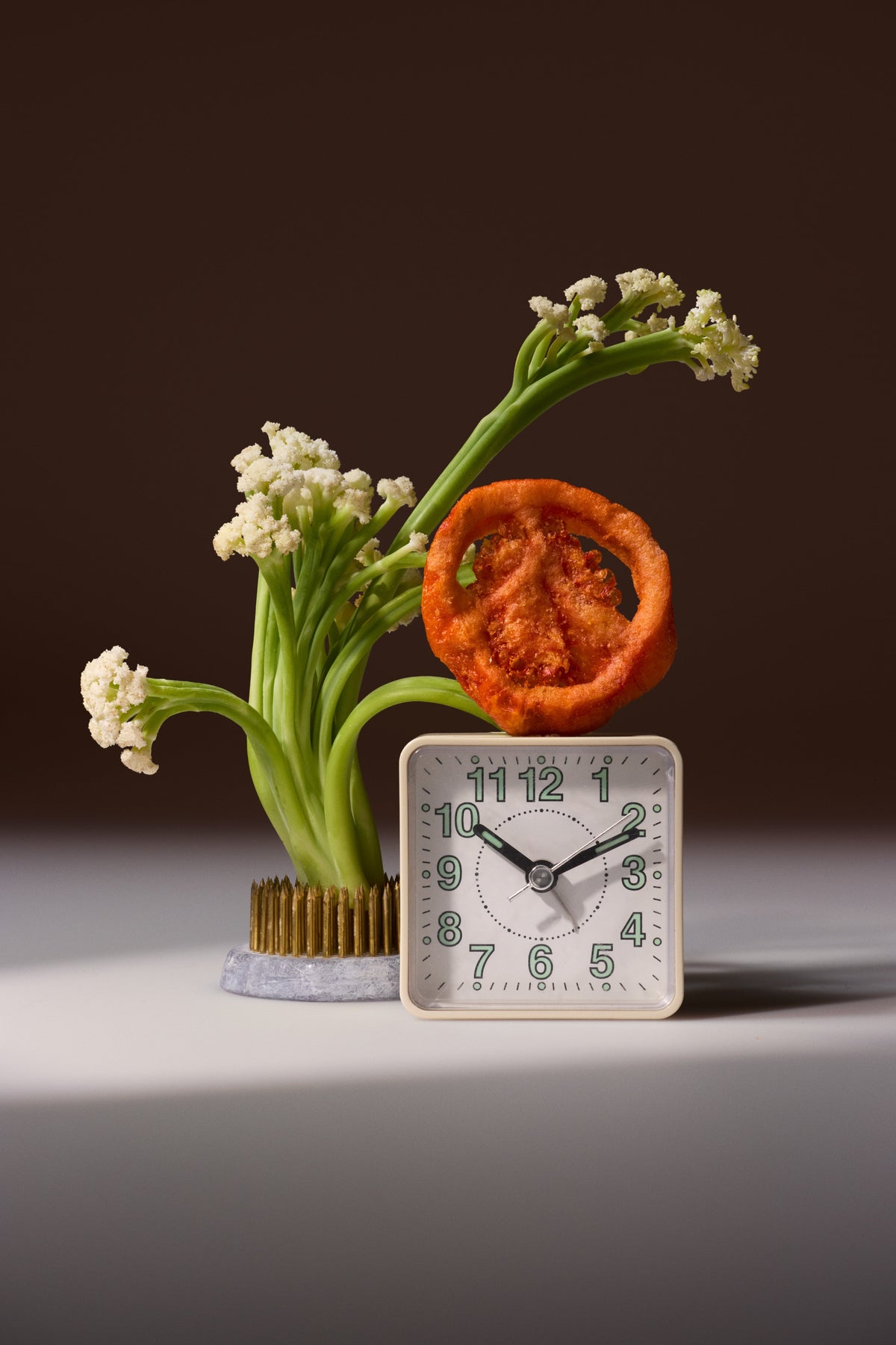 A square white alarm clock with black hands and green numbers sits on a light gray surface against a dark brown background, styled with a dried orange slice and white flowers in a gold-toned vase.