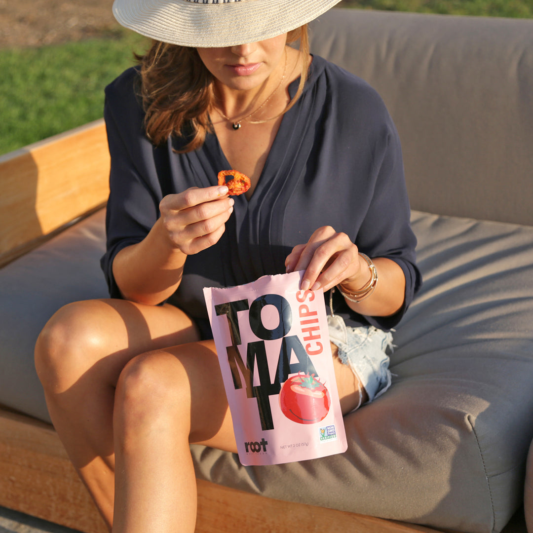 Lifestyle image of woman sitting outside on a couch, eating a Root Foods Tomato Chip out of a bag